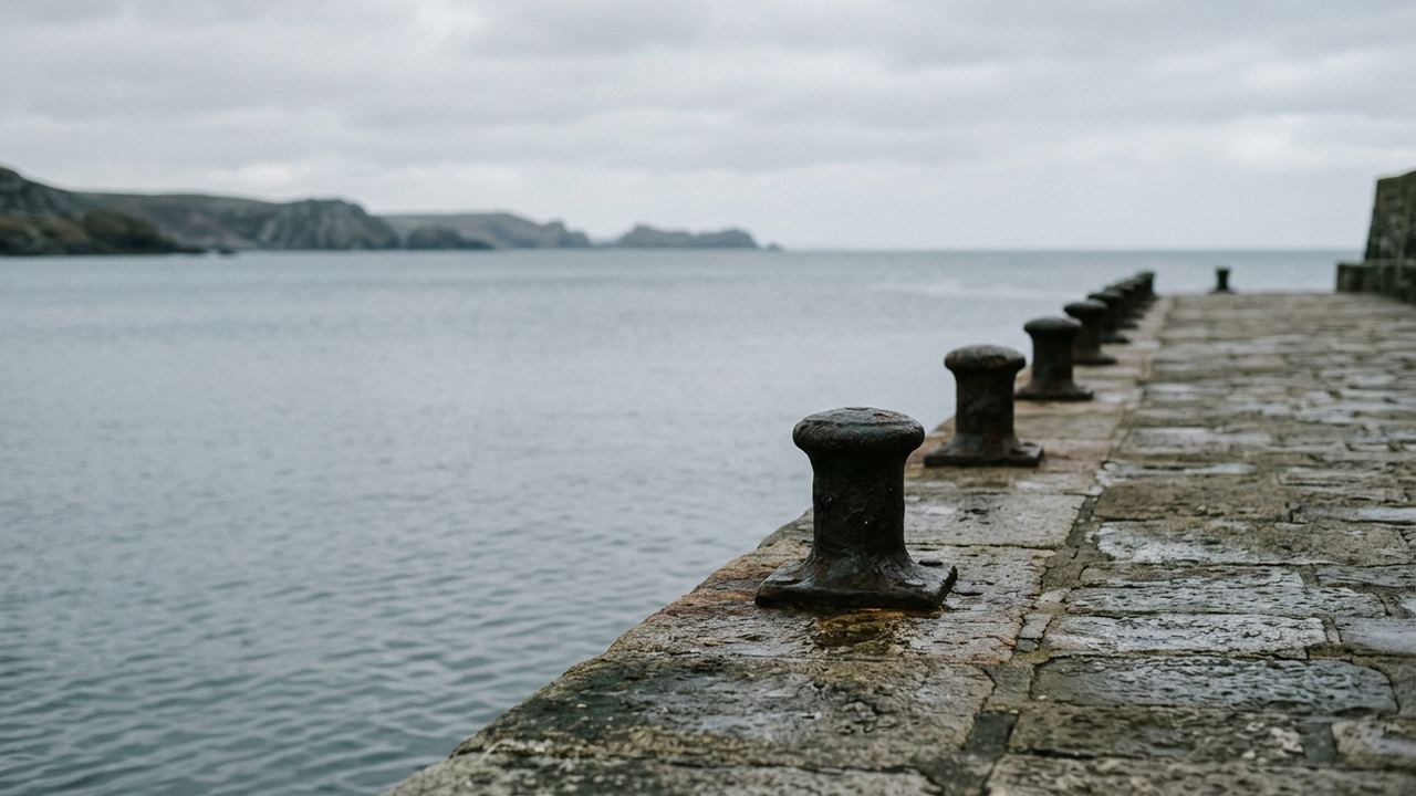 A quiet harbour wall with a receding line of iron bollards along it, calm grey water beyond, no boats